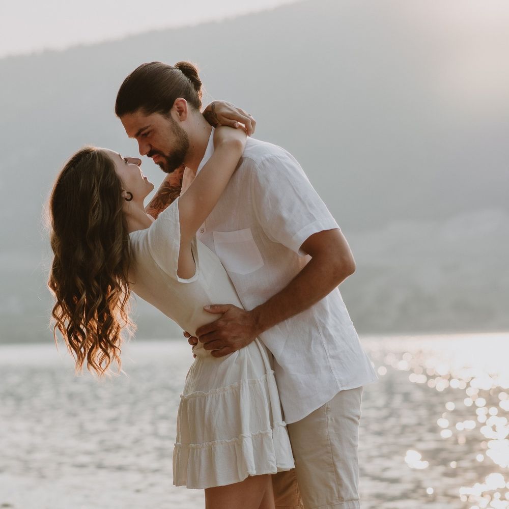 Couple sharing a romantic moment by a serene lake, dressed in white, embracing and smiling at golden hour.