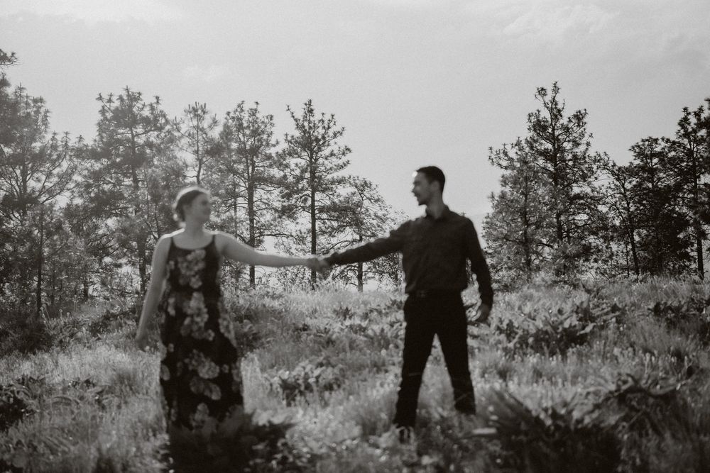 Black and white photo of a couple holding hands in a forest clearing, woman in floral dress, man in dark shirt.