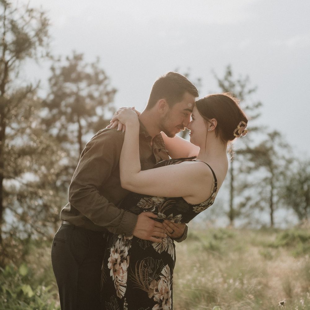 A couple embraces tenderly in a sunlit outdoor setting, touching foreheads romantically among trees and green grass.