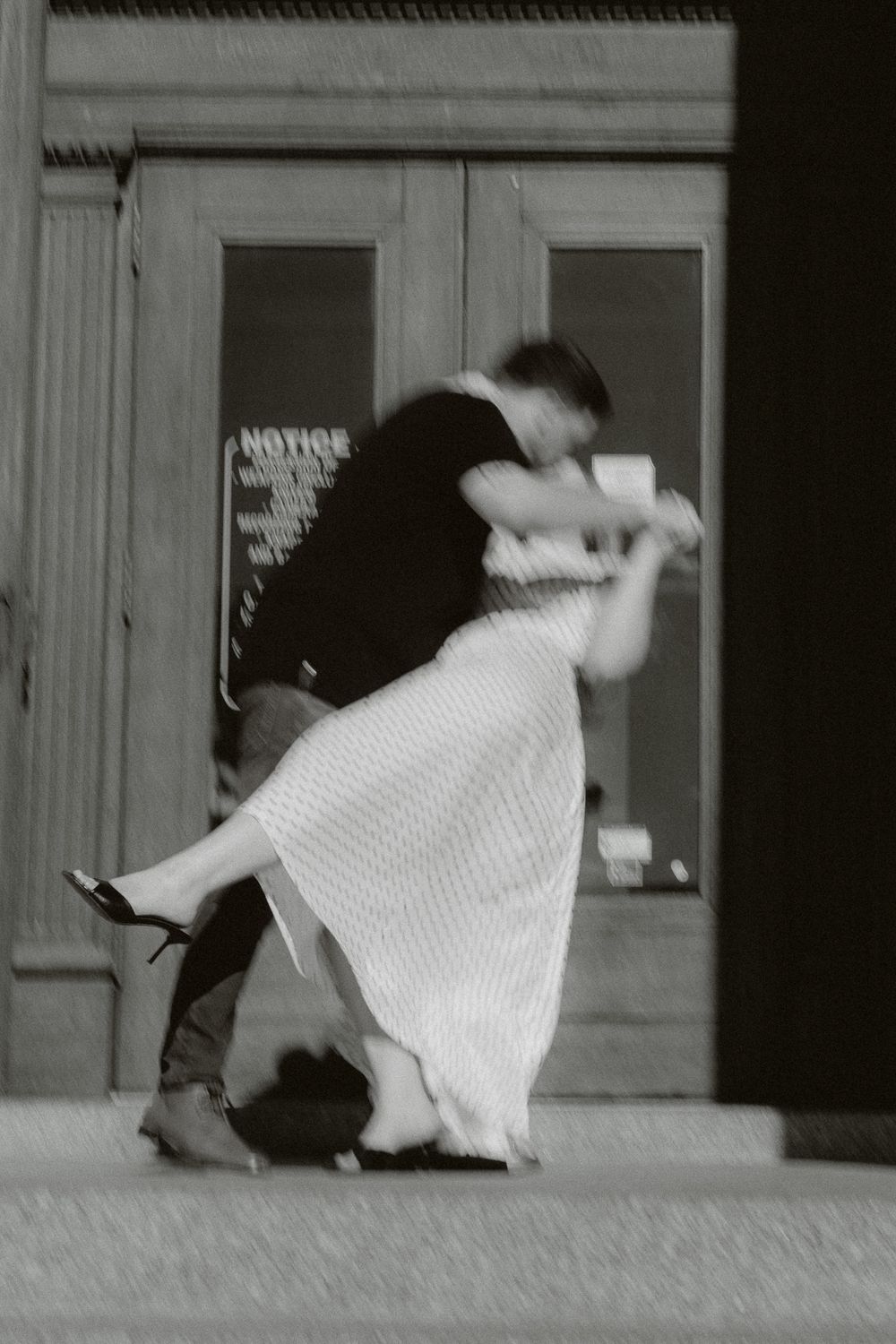 Black and white photo of a couple dancing and dipping on a sidewalk in front of a building with glass doors.