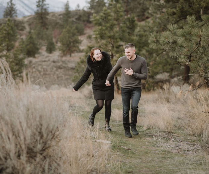 A happy couple laughs and walks together through a scenic outdoor trail surrounded by tall grass and pine trees.