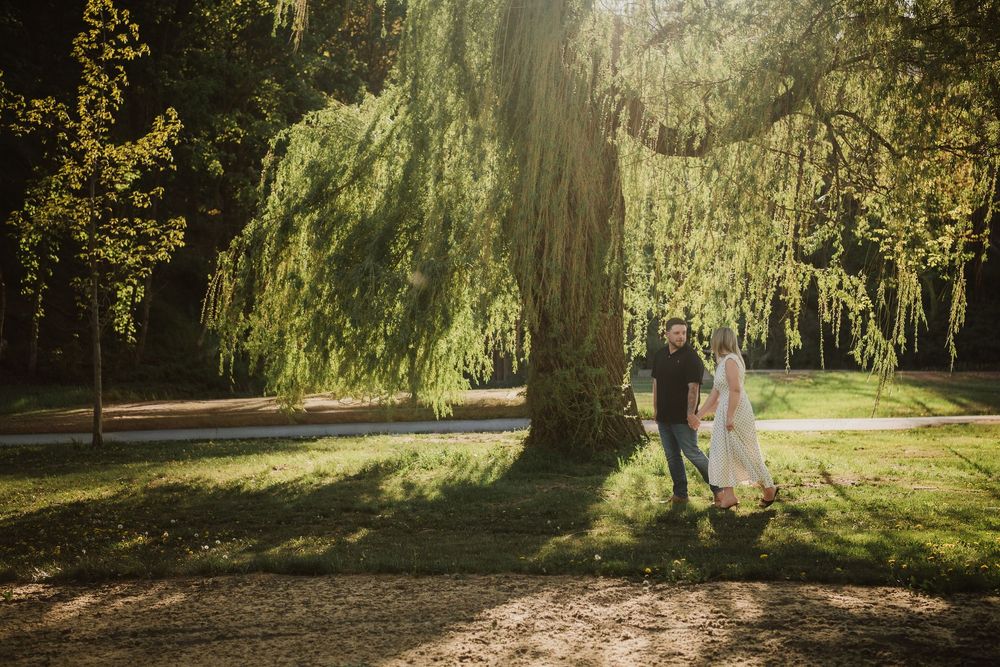 Couple holding hands under a large weeping willow tree in a sunlit park during golden hour.
