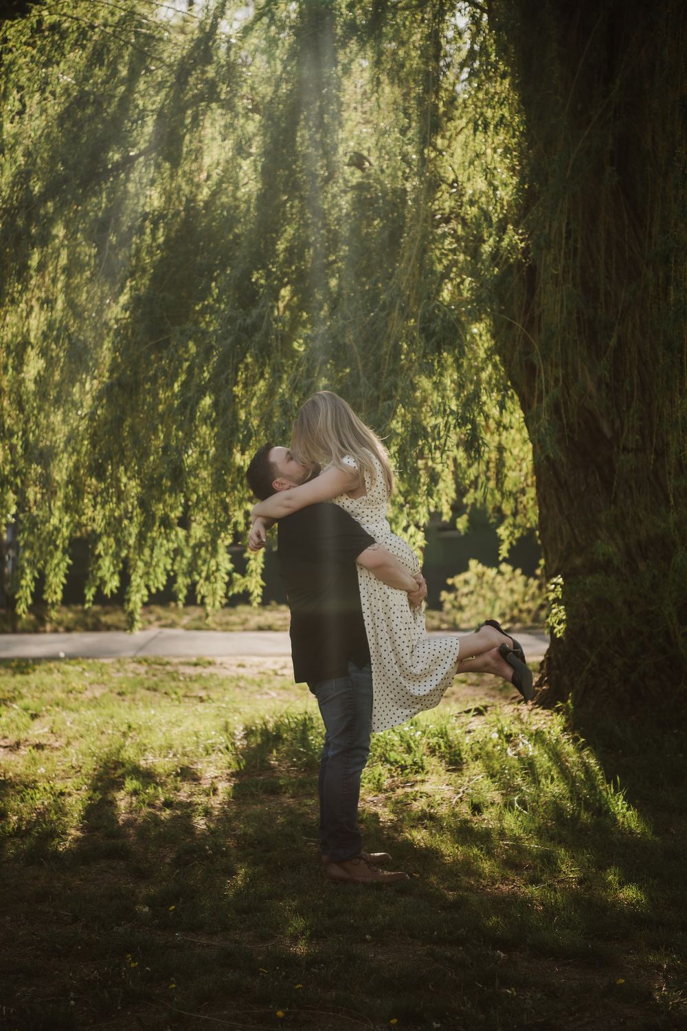 Couple embracing romantically under a weeping willow tree in a sunlit park, man lifting woman in a tender moment.