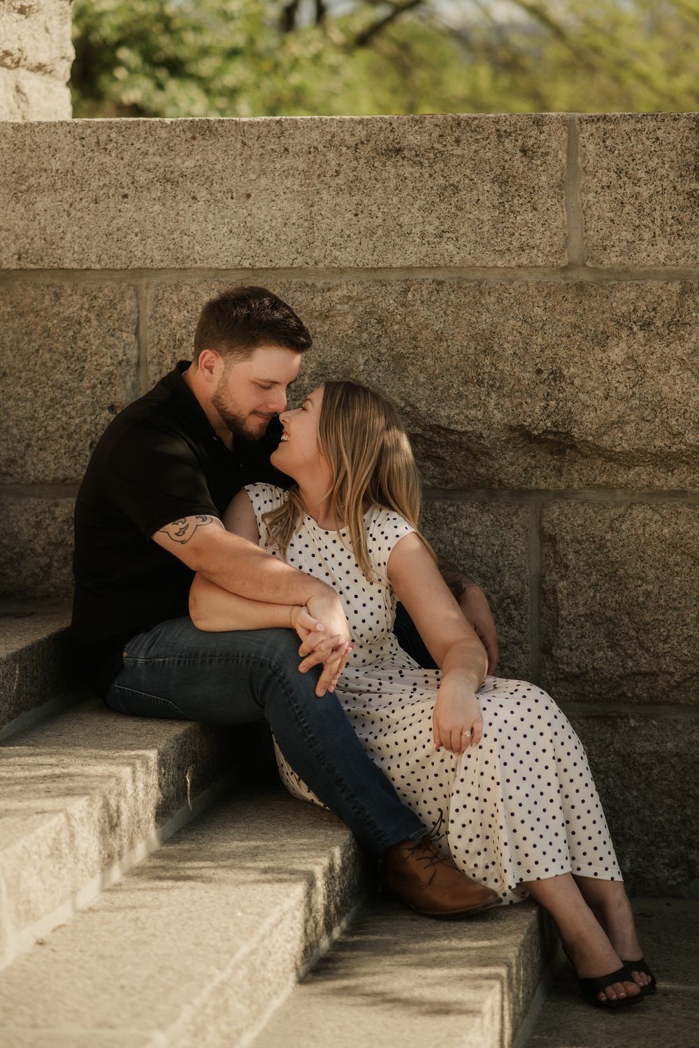 A couple shares a romantic moment sitting on stone steps, the woman in a polka dot dress leaning in for a kiss.