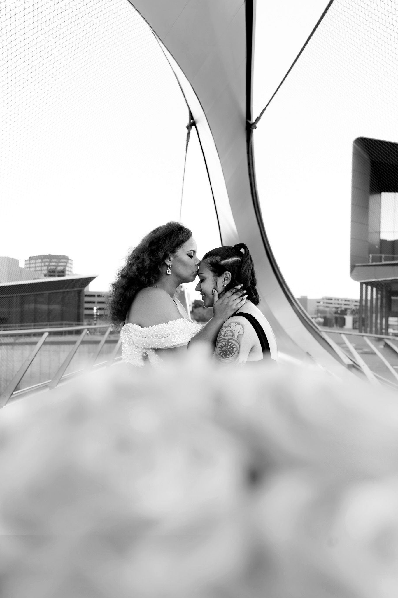 Black and white photo of two brides sharing a kiss on a modern bridge, with city buildings in the background.