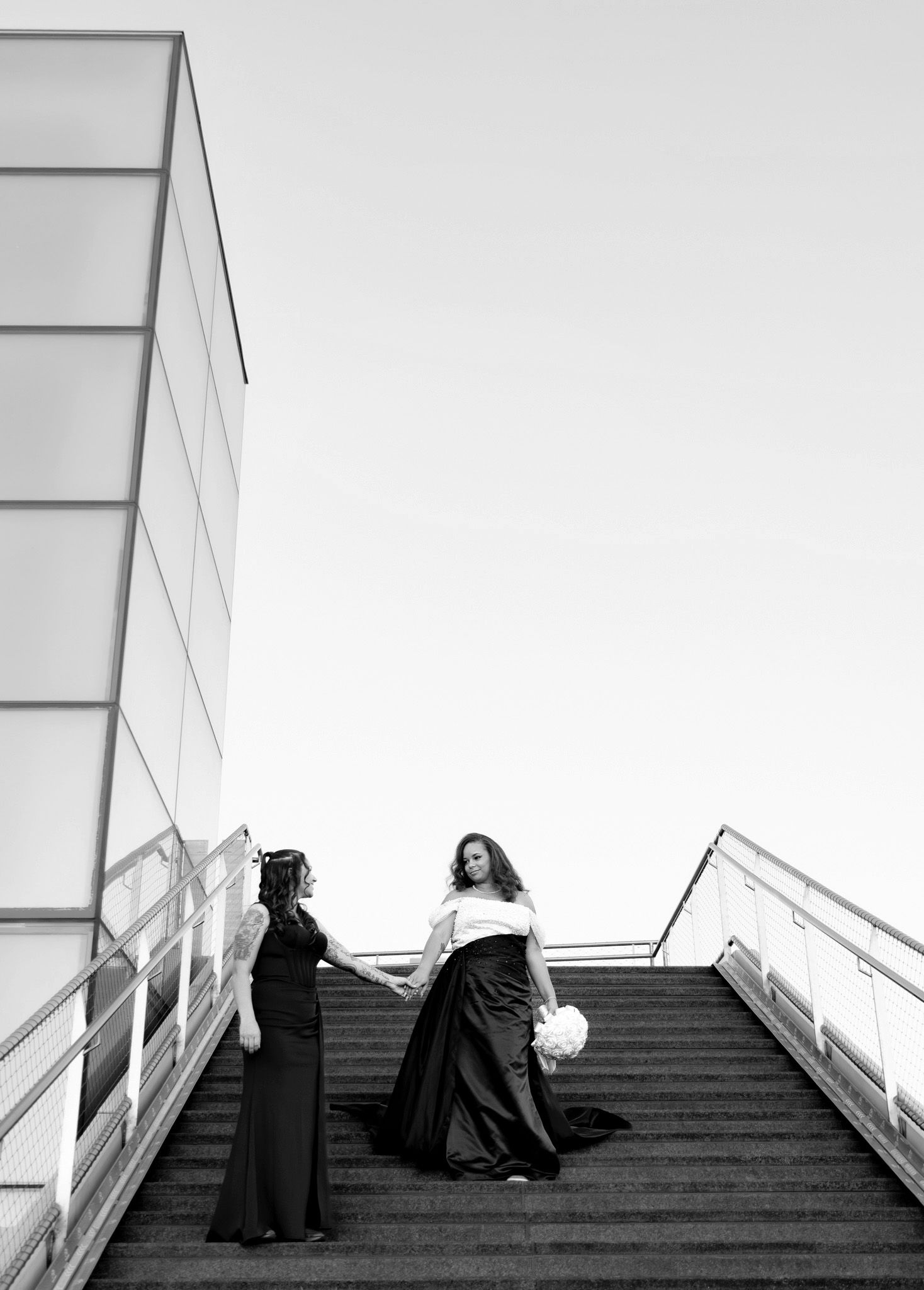 Two brides in black and white gowns hold hands on outdoor stairs in a romantic black and white wedding photo.