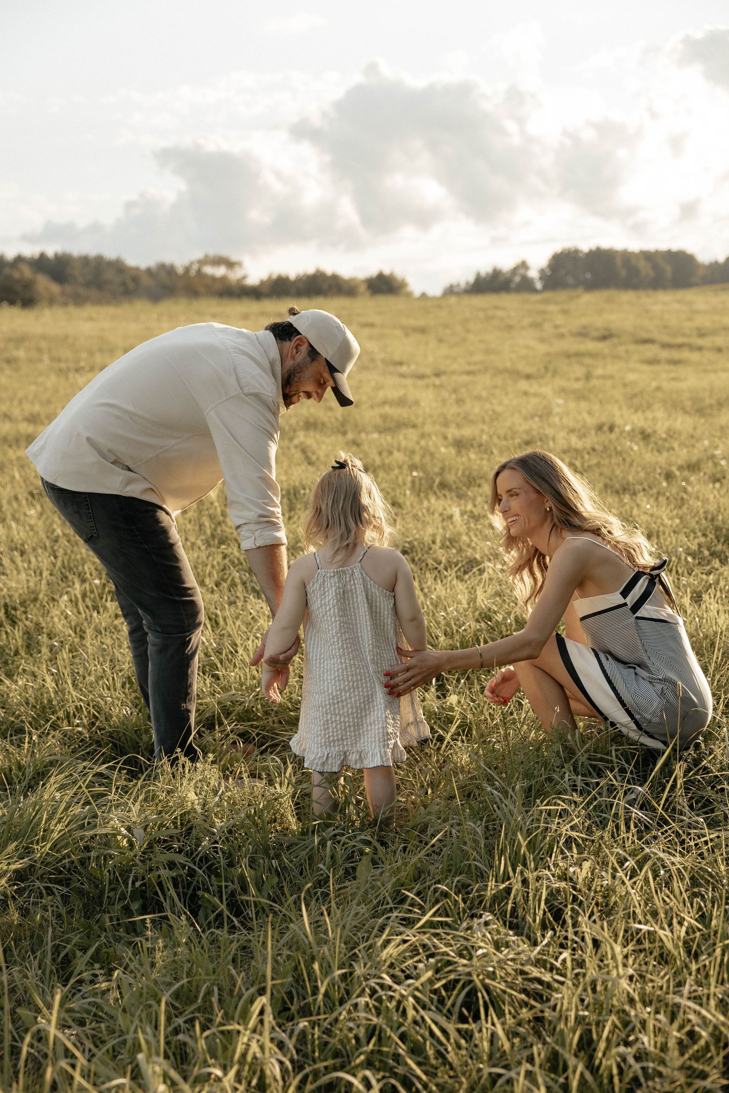 A young family with a toddler girl enjoys a sunny day in a lush green field, with trees and clouds in the background.