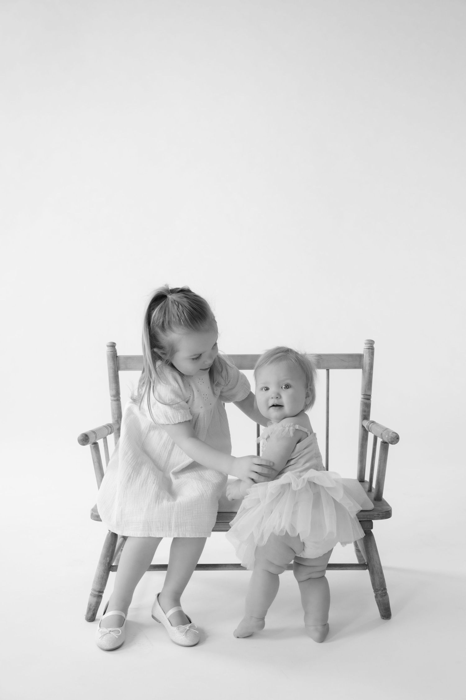 Toddler helps her baby sister stand near a bench during in-studio family photos in Stillwater, MN.