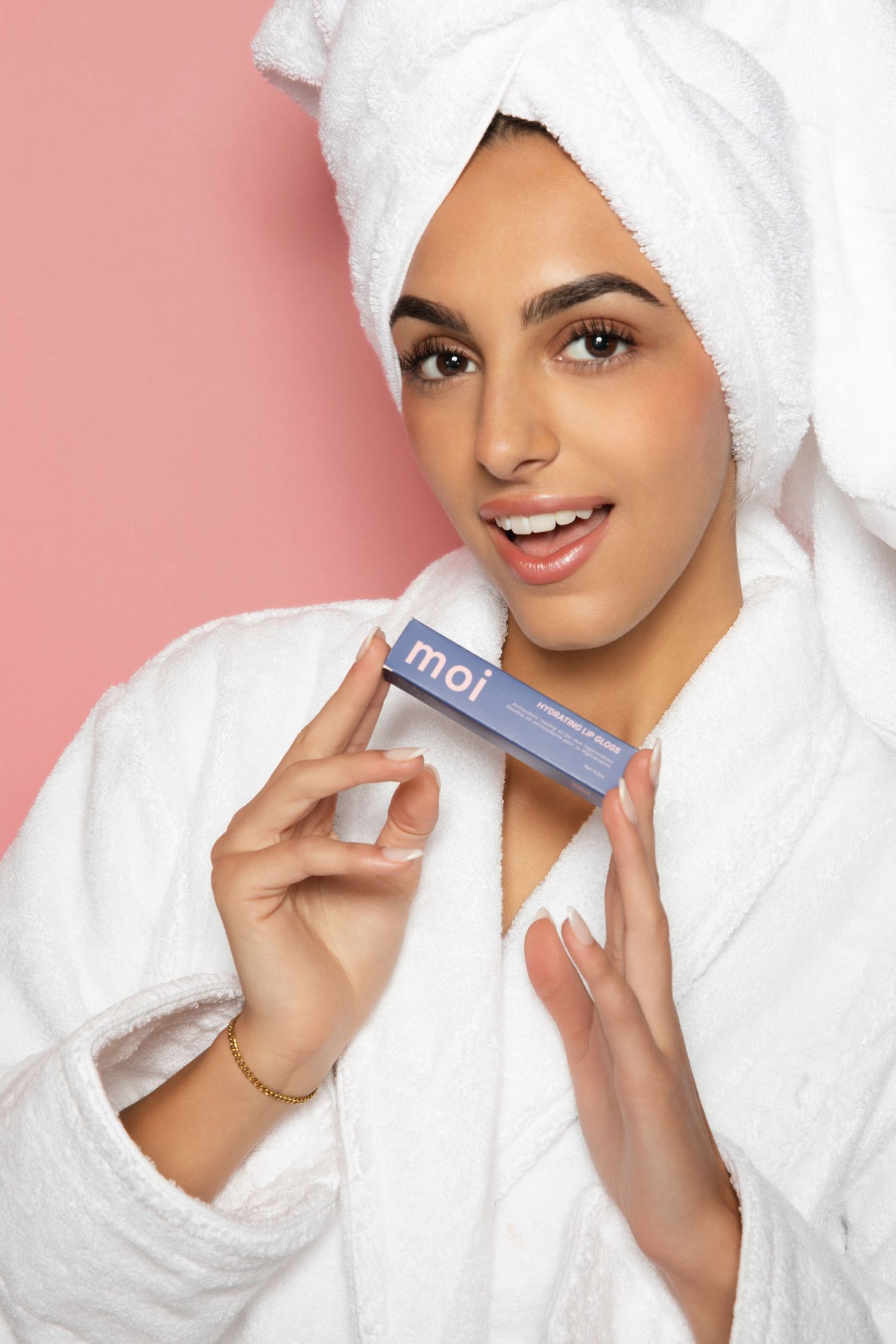 Woman in white bathrobe and towel turban holding Moi skincare product, smiling against pink background.