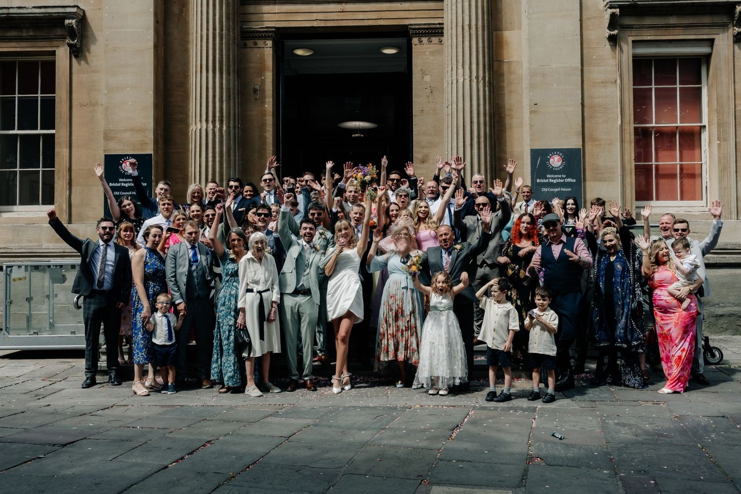 Large wedding group photo outside a grand stone building, guests cheering with raised arms in festive attire.