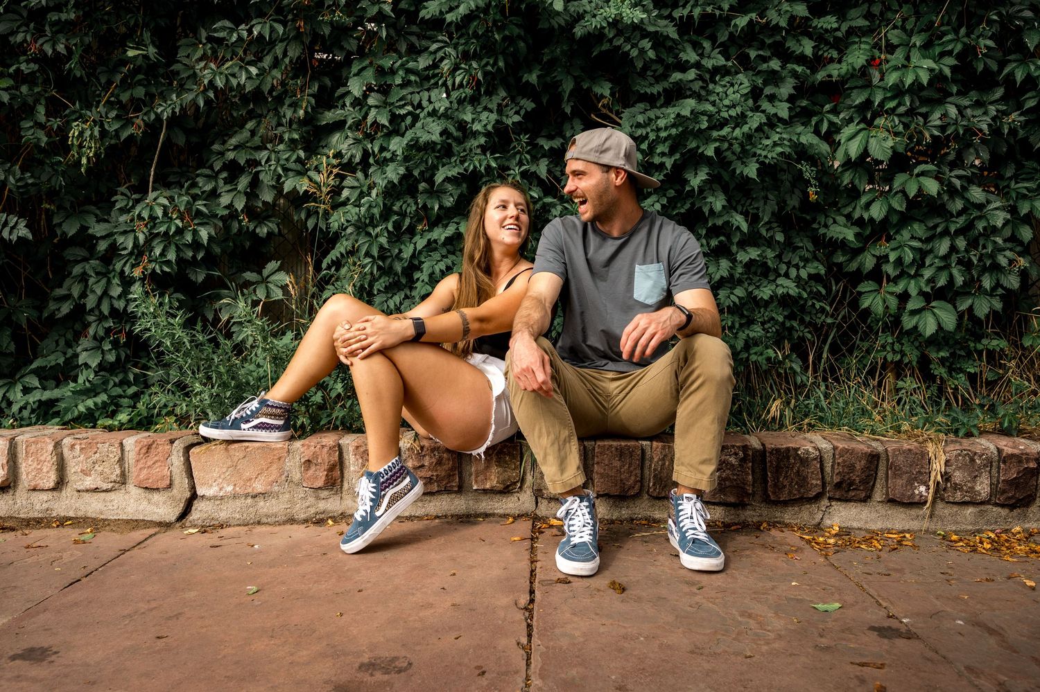 A smiling couple sits on a brick wall surrounded by lush green foliage, both wearing casual outfits and sneakers.