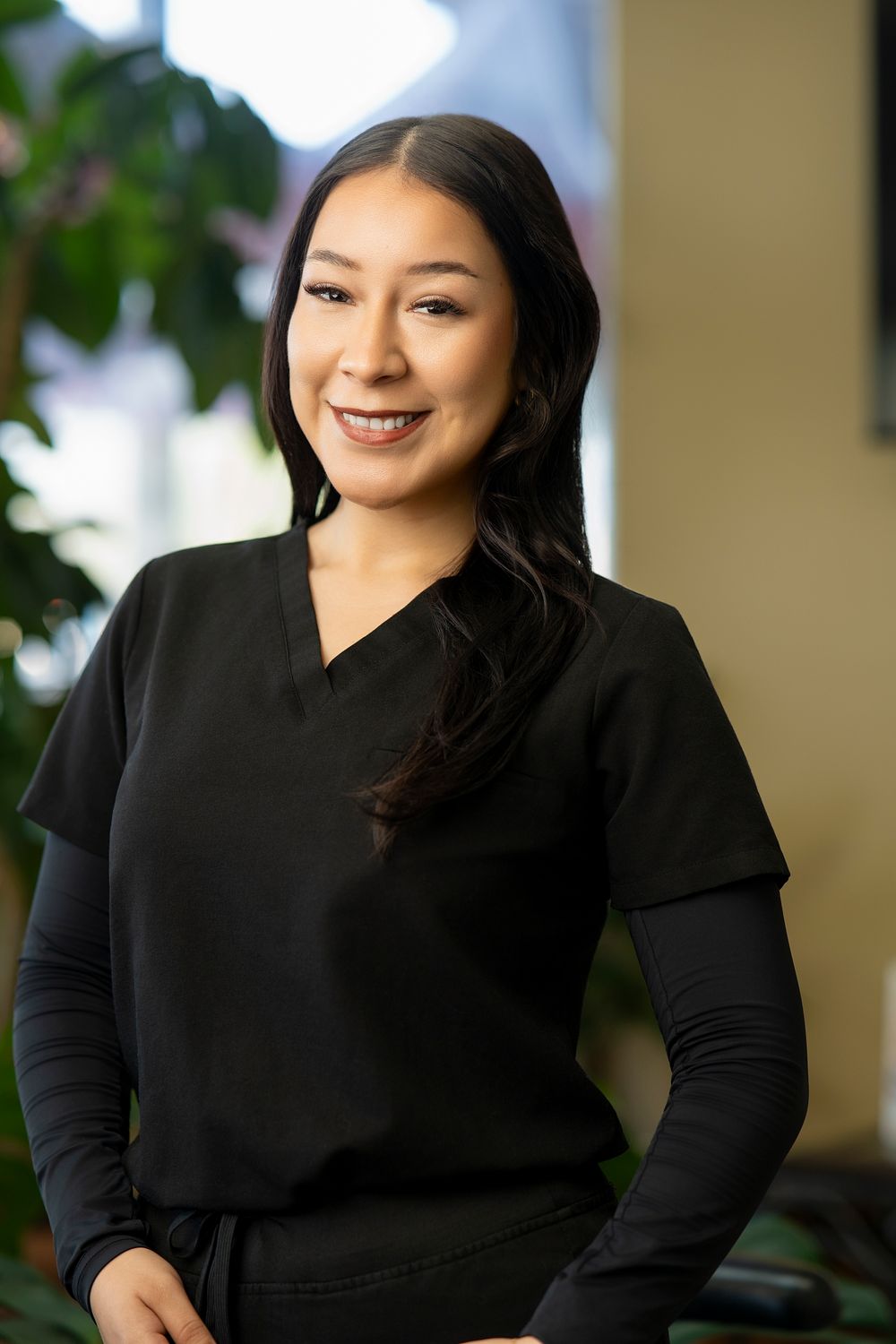 Smiling young woman in black scrubs stands confidently in a modern medical office with green plants in background.