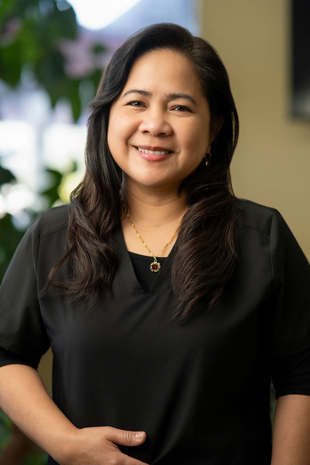 Smiling woman with dark hair wearing a black blouse and gold necklace posing indoors with greenery in background.