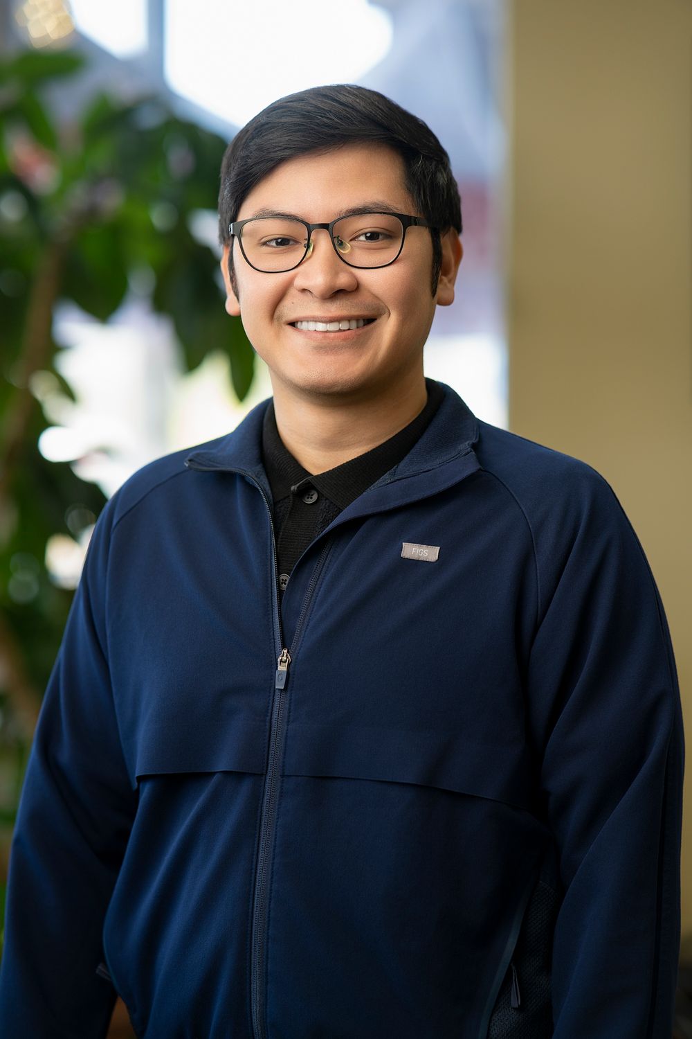 Smiling young man wearing glasses and a navy blue zip-up jacket, standing indoors with green plants in the background.