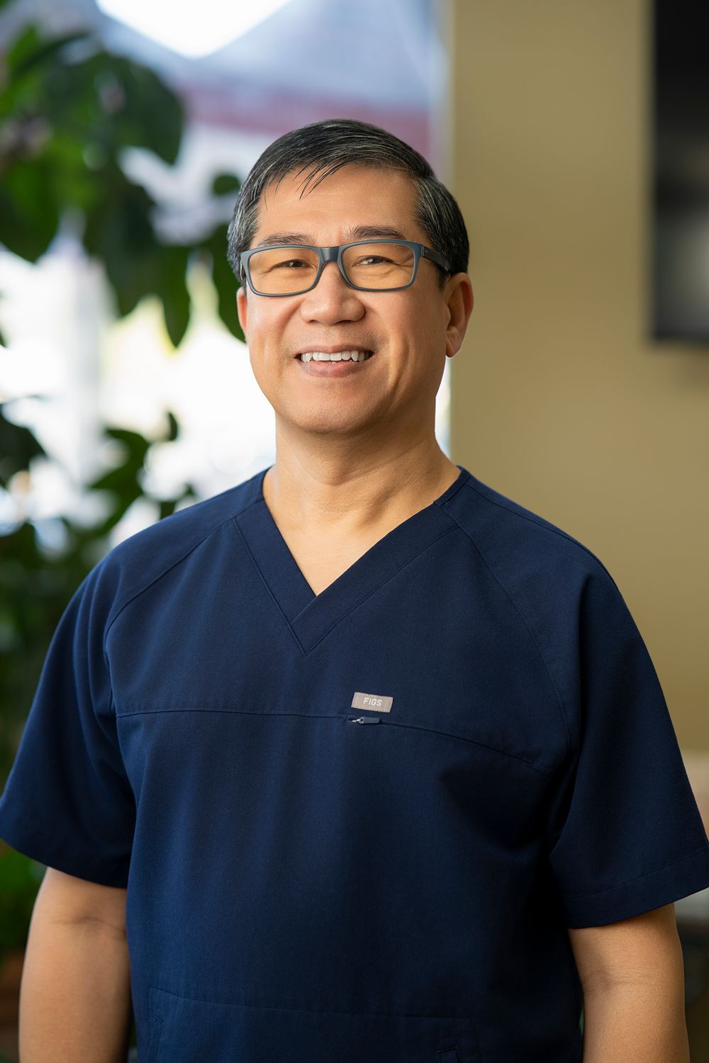 Smiling male healthcare professional in navy blue scrubs and glasses posing outdoors with green foliage background.