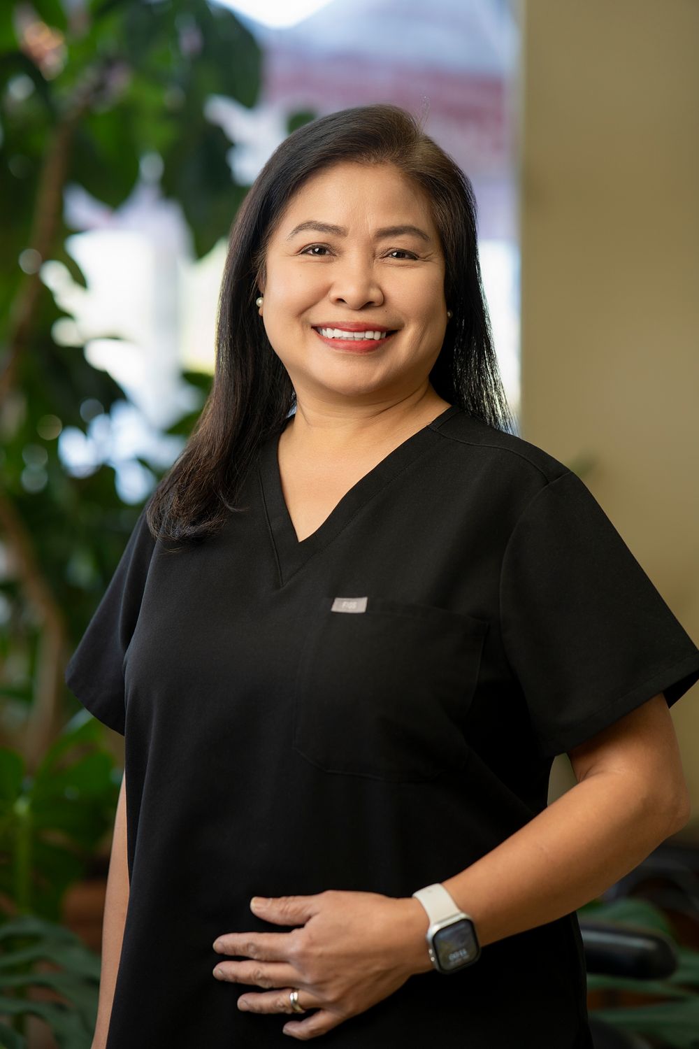 Smiling female healthcare professional in black scrubs poses confidently indoors with lush green plants in background.