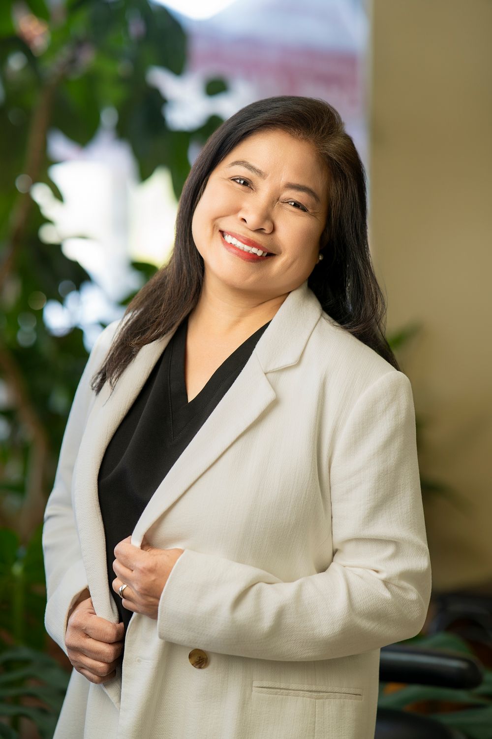 Smiling Asian businesswoman in white blazer and black top posing confidently indoors with green plants in background.