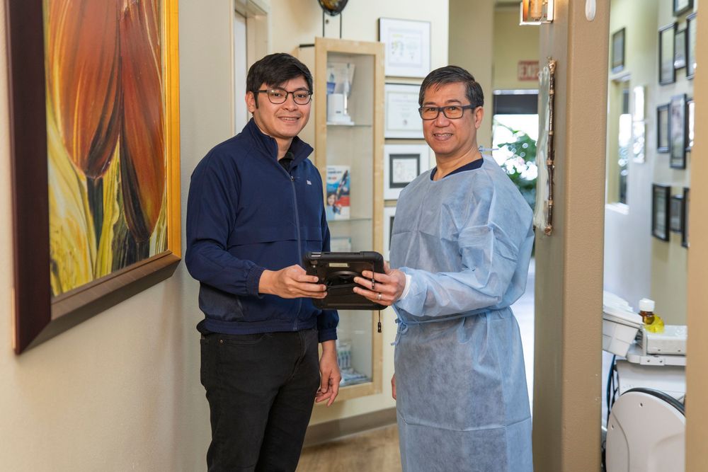 Two men in a dental office smiling and holding coffee cups, one wearing a blue shirt and the other in a medical gown.