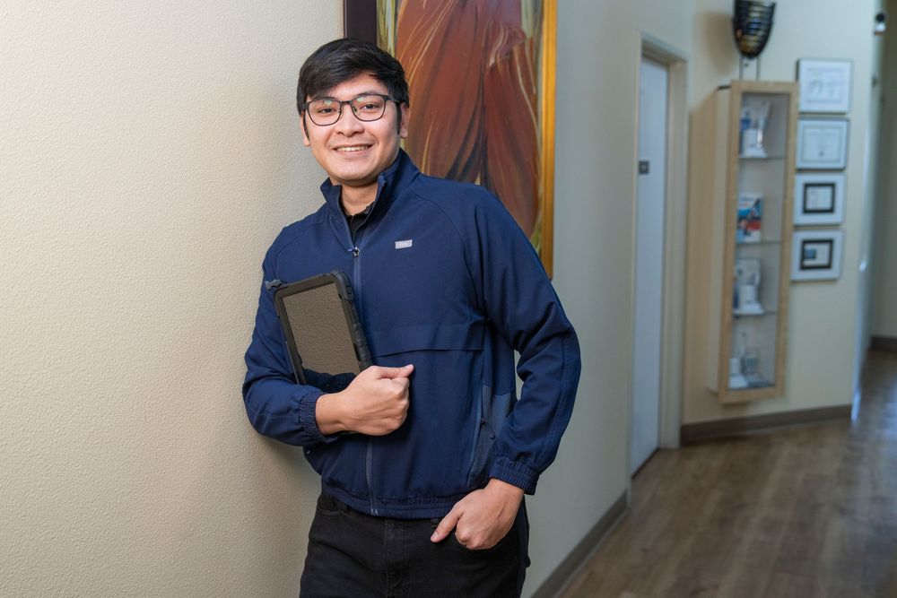 Smiling Asian man in navy blue jacket holding a tablet, standing in a modern office hallway.