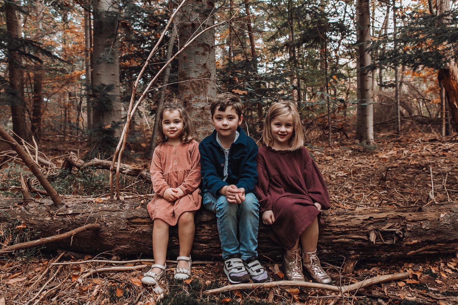 Three children sitting on a fallen log in an autumn forest, wearing cozy fall outfits among colorful leaves in Amsterdamsebos