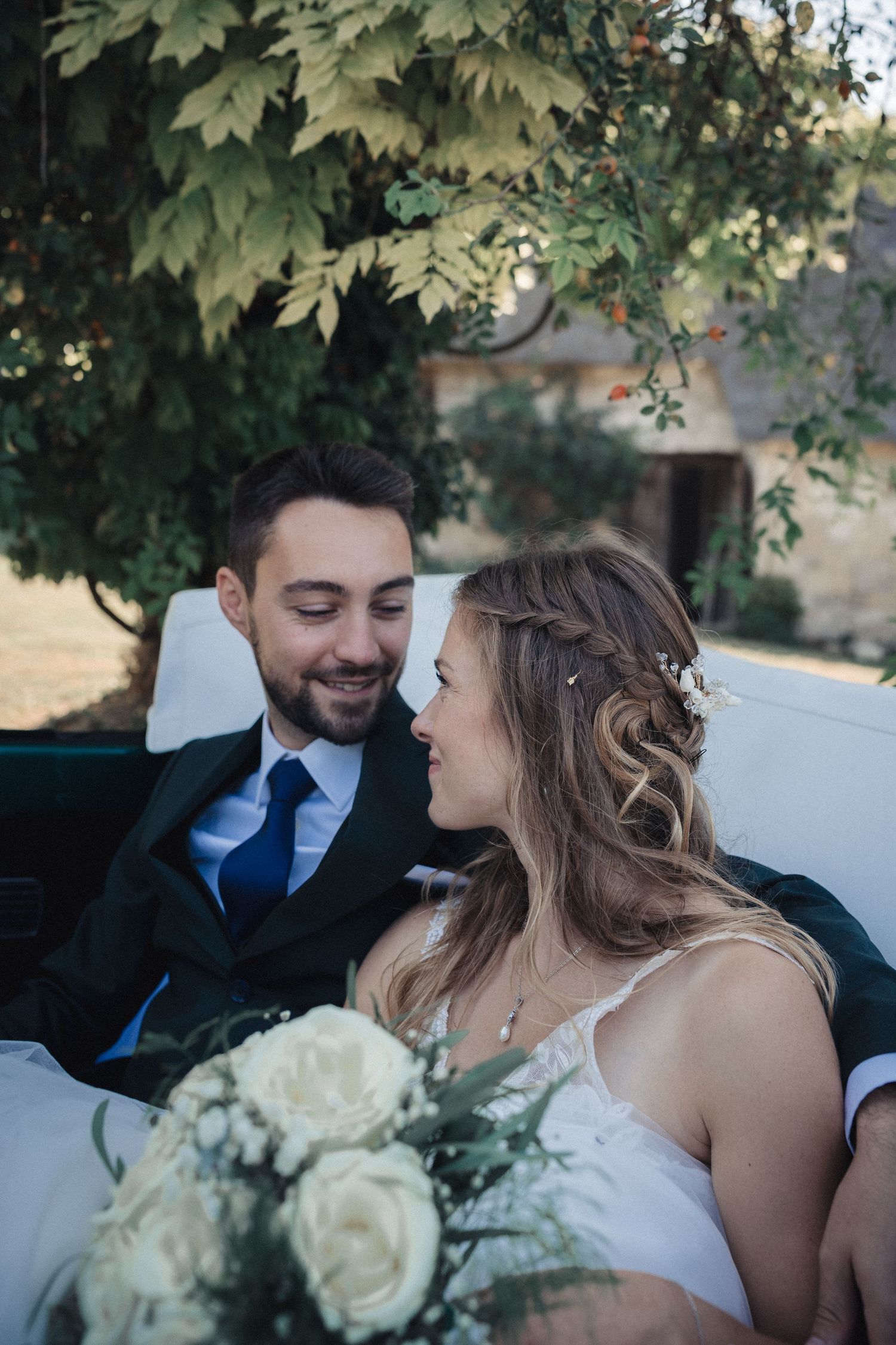 A romantic wedding couple embraces in a wooded outdoor setting with guests in the background.