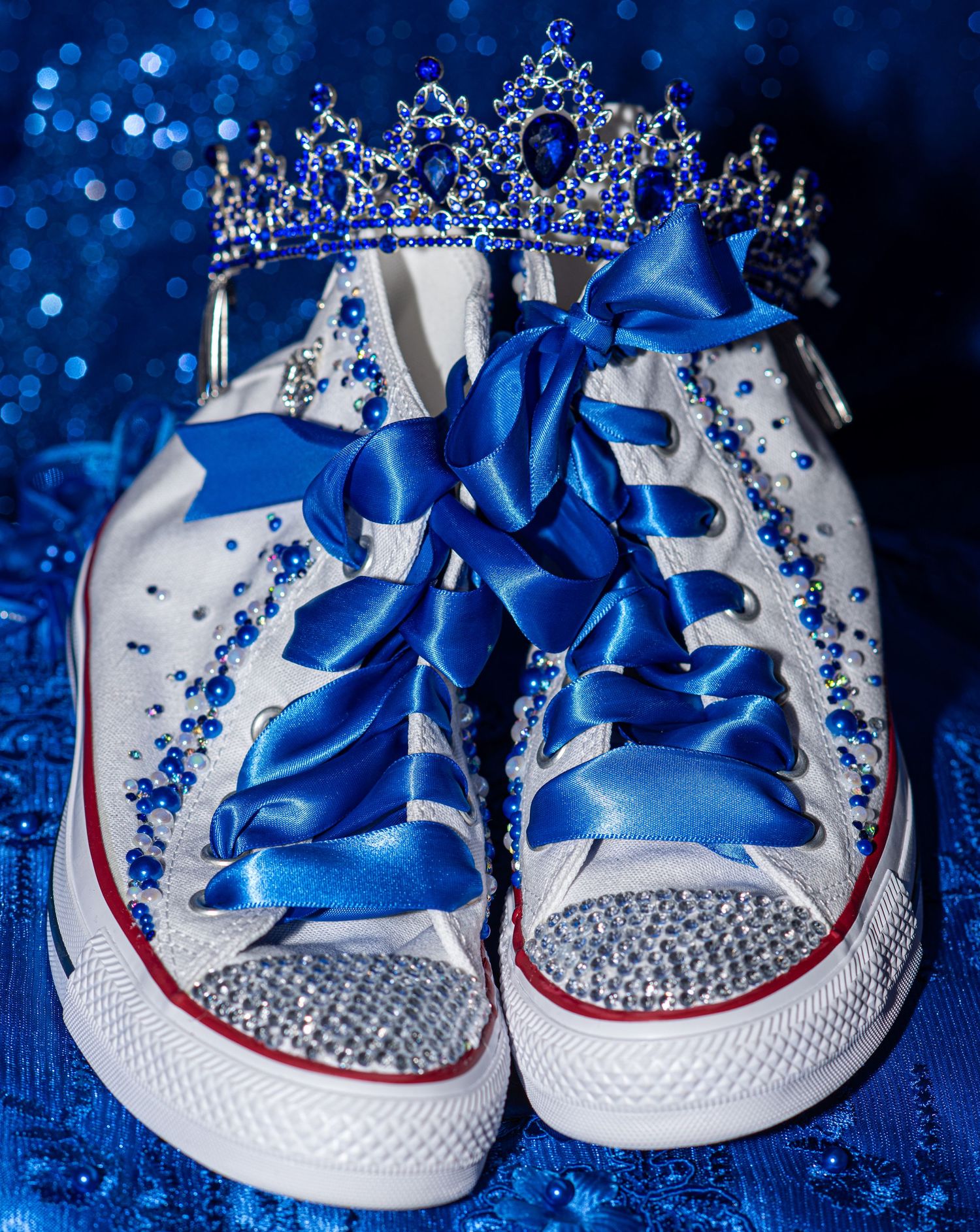 Rhinestone-decorated white Converse sneakers with blue satin laces and tiara for quinceañera celebration.