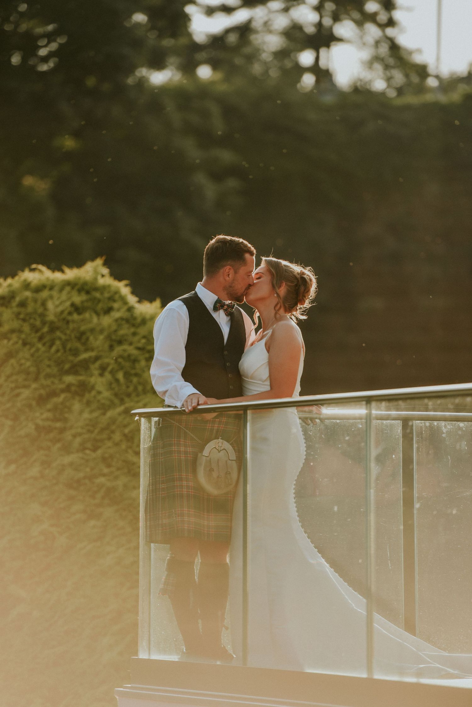 Bride and groom sharing a romantic kiss on a balcony at golden hour, groom wearing a traditional Scottish kilt.