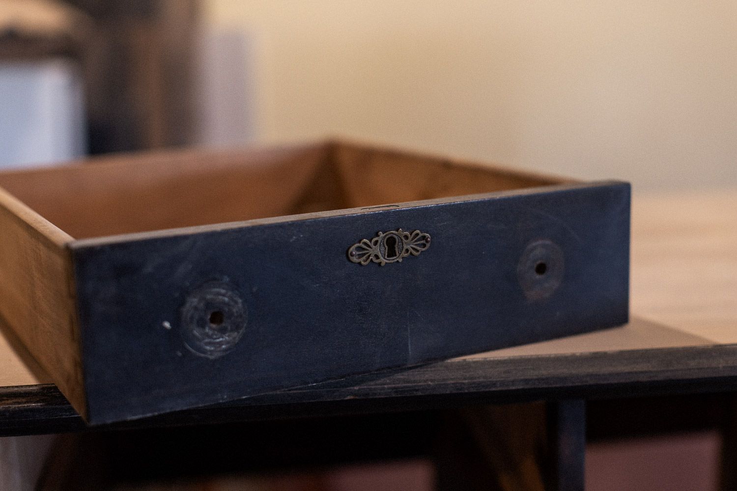 Antique wooden drawer with dark metal front panel, decorative keyhole escutcheon, and round knobs on a rustic table.