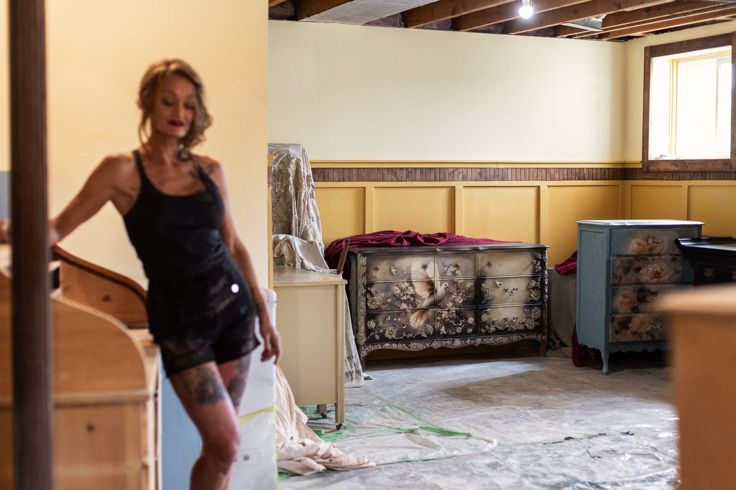 Woman in overalls stands in a room under renovation with drop cloths and artwork visible.