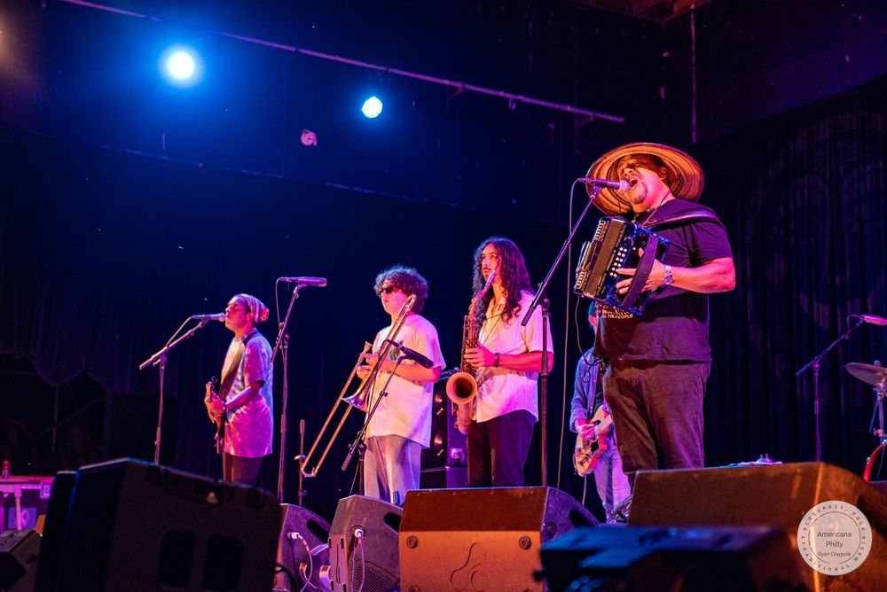 Live band performing on stage with guitar, trombone, saxophone, and accordion under blue stage lights.