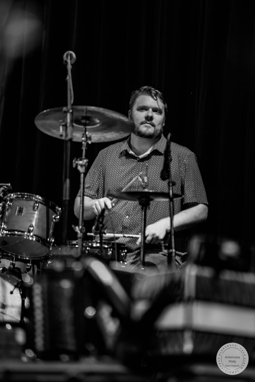 Black and white photo of a male drummer performing on stage with cymbals and drum kit visible in background.