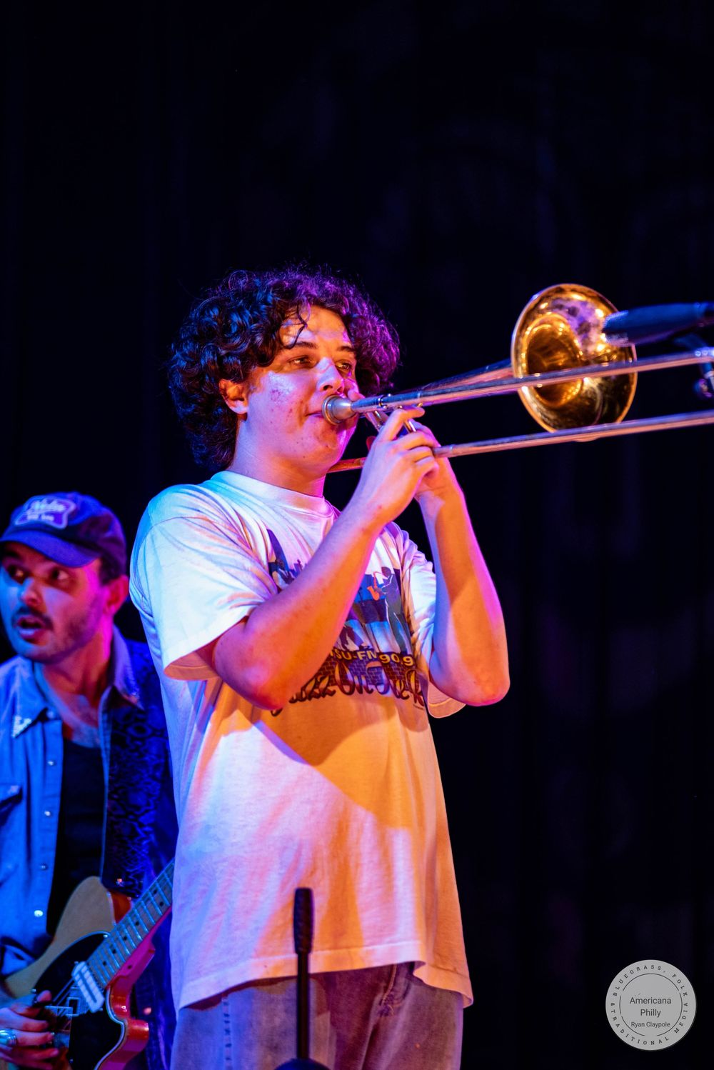 Young musician plays trombone on stage under colorful lights, with a guitarist visible in the background.