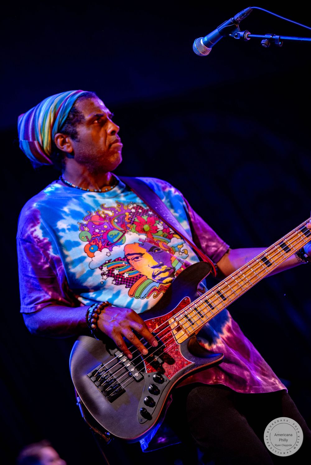 Musician in tie-dye shirt plays bass guitar on stage under colorful lights.