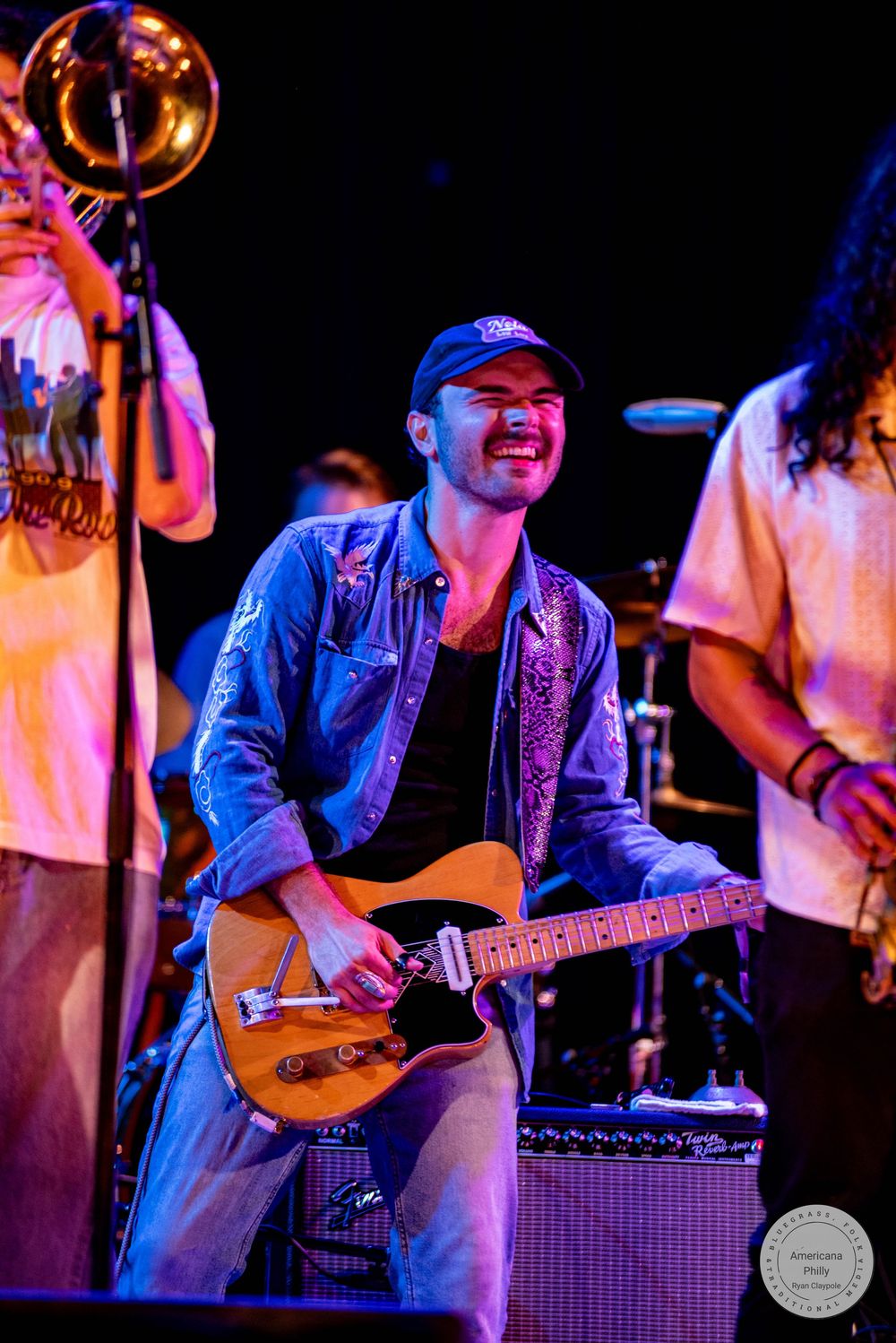 Smiling male guitarist in denim jacket and blue cap performs on stage with band members and trombone player visible.