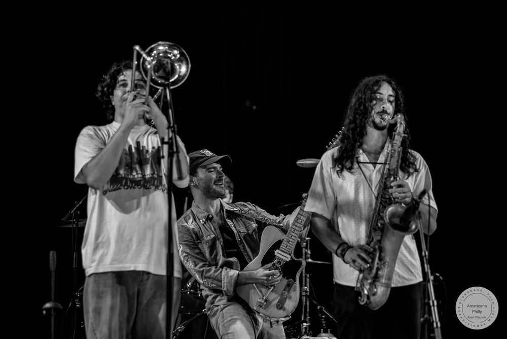 Black and white photo of three musicians performing live, playing trumpet, guitar, and saxophone on stage.