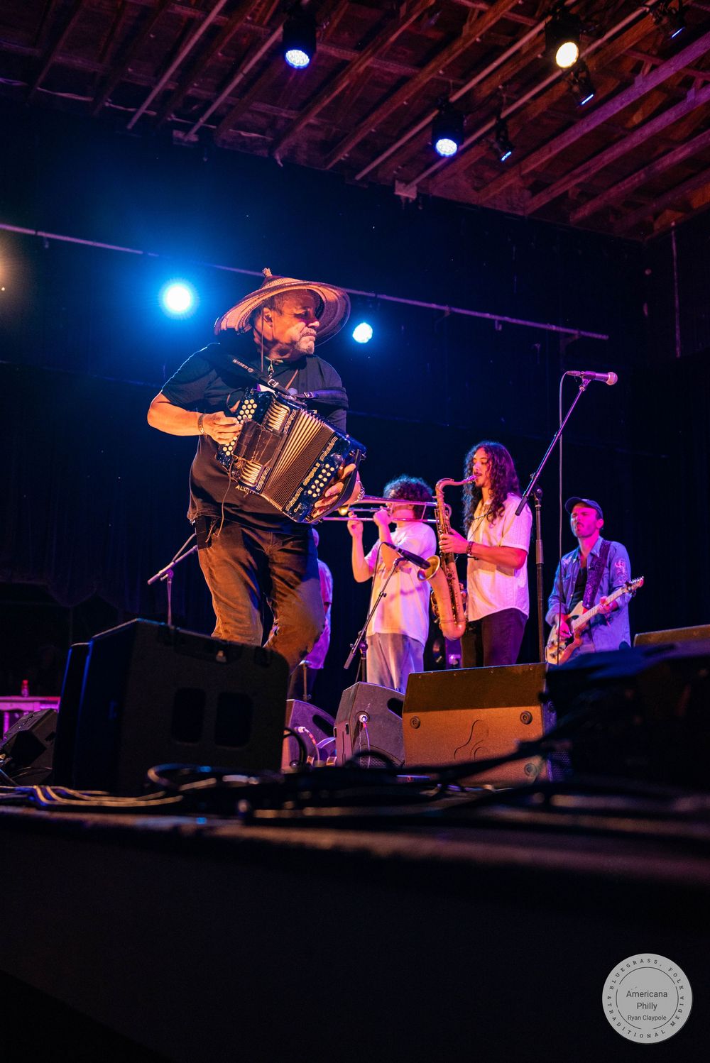 Musician playing accordion on stage wearing a wide-brim hat with band members playing flute, saxophone, and guitar behind him.