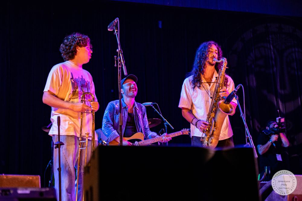 Three musicians perform on stage, two standing and one seated with a guitar, in a warmly lit venue.