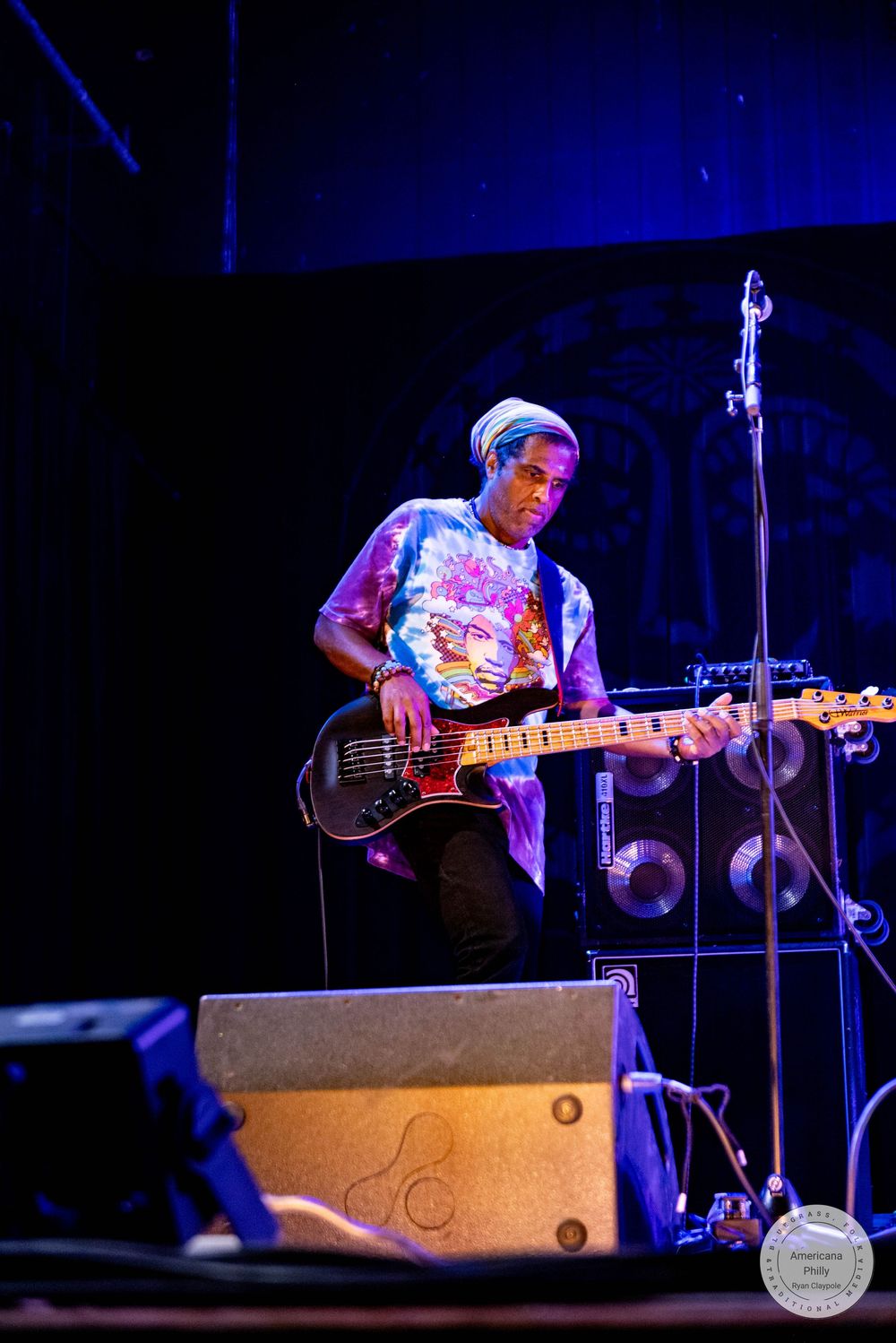 Musician playing electric bass guitar on stage wearing a tie-dye shirt under blue stage lighting.