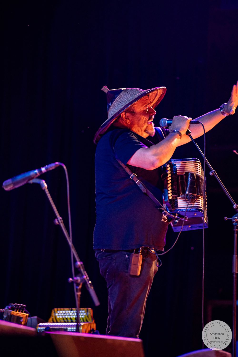 Man in wide-brim hat playing accordion and singing into microphone on stage during live music performance.