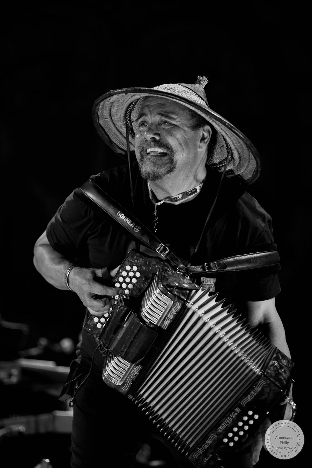 Black and white photo of a smiling man playing accordion while wearing a wide-brimmed straw hat on stage.