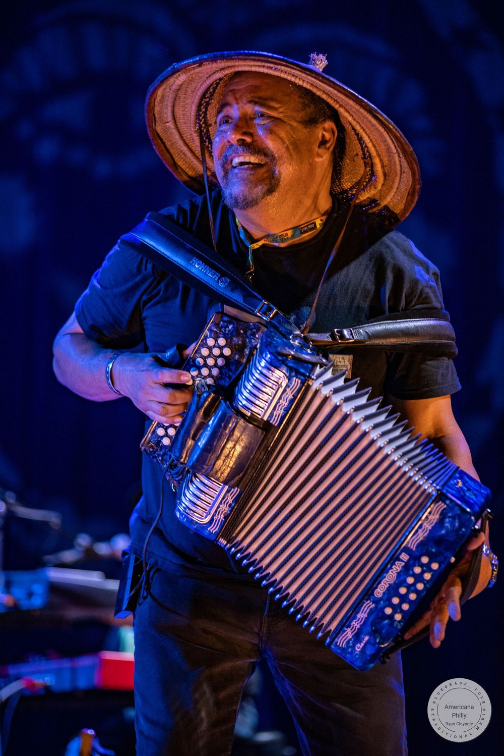 Smiling musician playing a blue accordion on stage, wearing a straw hat and black shirt under blue stage lighting.