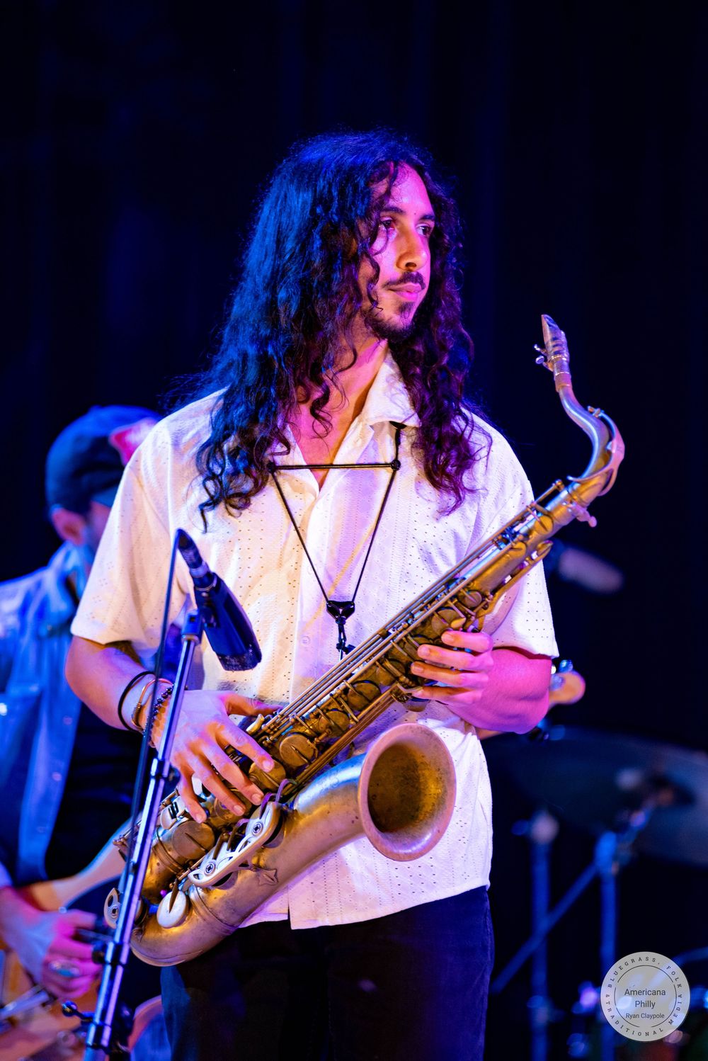 Young male musician with curly hair plays saxophone on stage in white shirt under blue stage lighting.