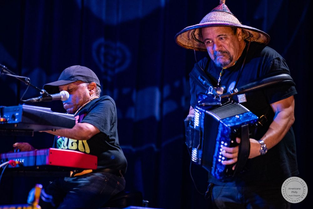 Two musicians perform on stage, one playing keyboard and one playing accordion wearing a straw hat.