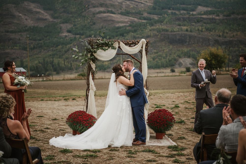 Bride and groom share first kiss at outdoor mountain wedding ceremony with red mums and floral arch backdrop.