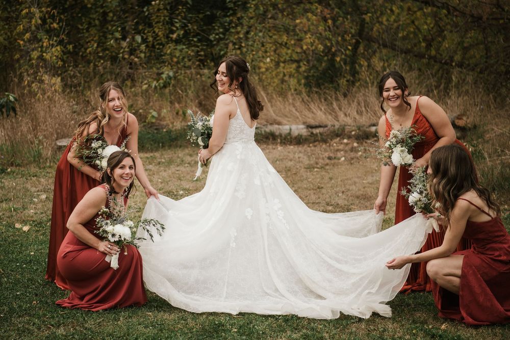 Bride in white gown with bridesmaids in red dresses arranging her train outdoors in a fall setting.