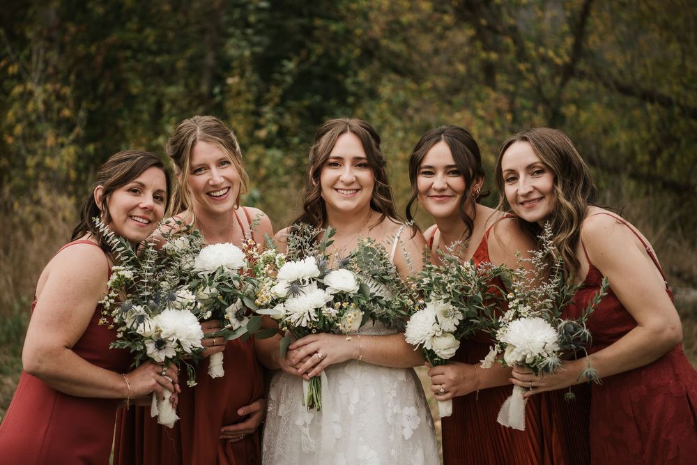 Bride in white lace gown with four bridesmaids in rust dresses holding white floral bouquets outdoors.