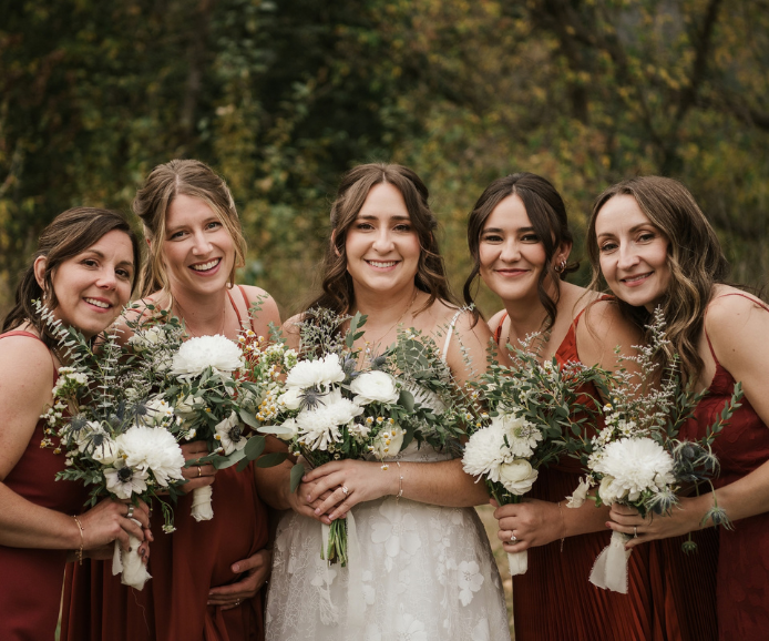 Bride in white gown with four bridesmaids in burgundy dresses holding white floral bouquets outdoors.