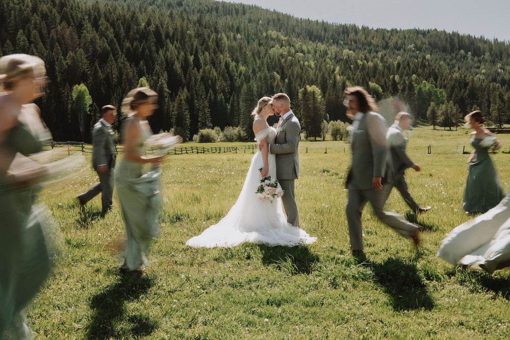 A bride and groom share a kiss in a sunlit meadow surrounded by their wedding party in sage green attire with forest backdrop.