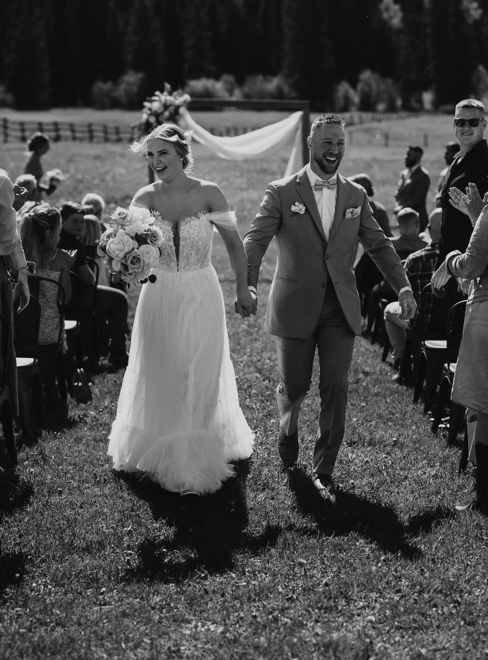 Joyful newlyweds walk down the aisle after outdoor wedding ceremony, bride in strapless gown holding bouquet, groom in suit.