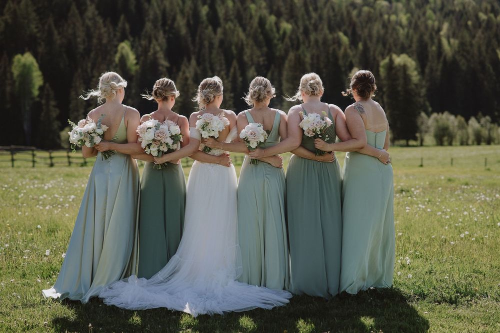 Bride and bridesmaids in sage green gowns holding white bouquets, standing in a sunlit mountain meadow.